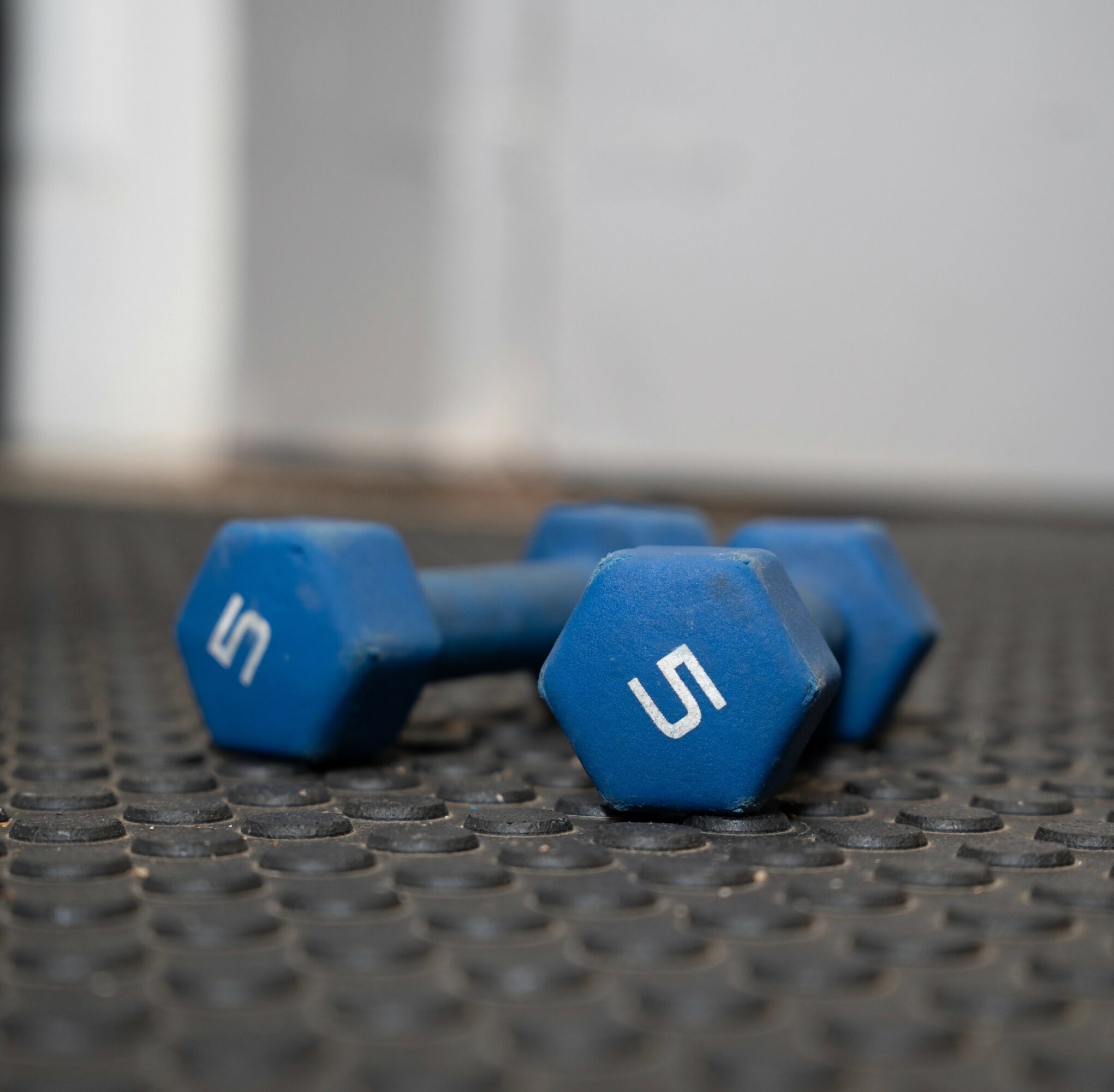 Two blue 5-pound dumbbells on a textured black surface, with a blurred background.