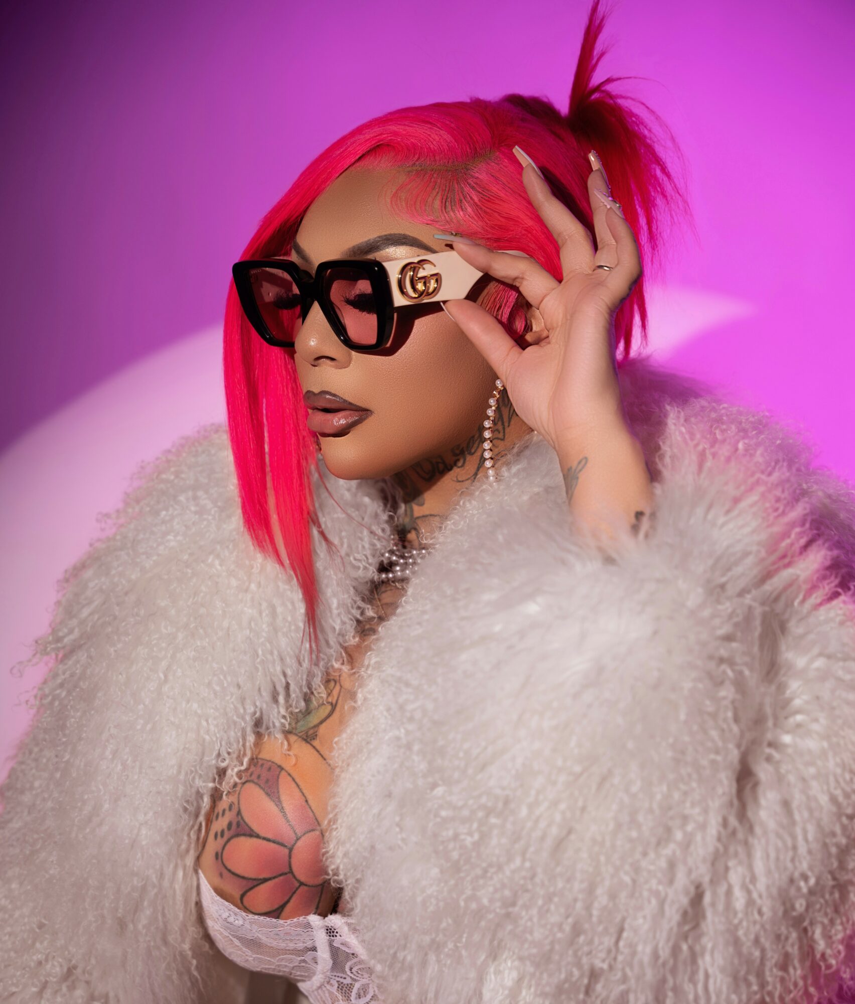 Woman with pink hair, large sunglasses, and a fluffy coat, holding her glasses with a pink and purple background.