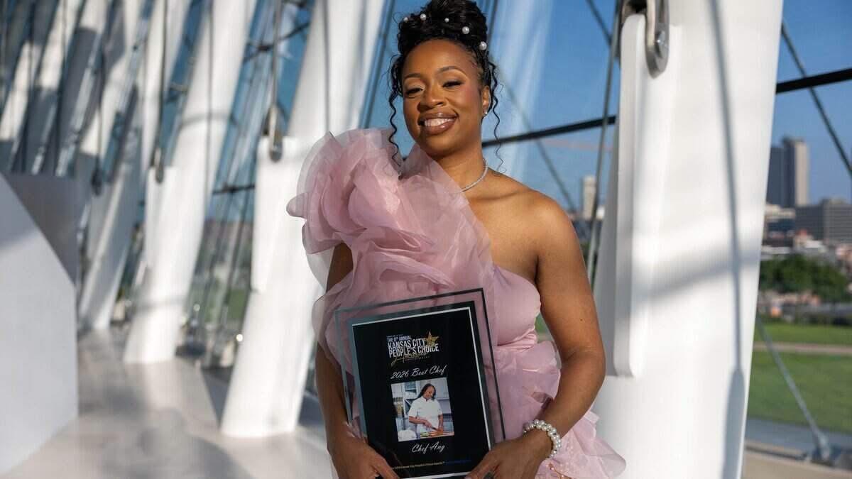 Woman in pink dress holding a framed award or certificate, smiling outdoors with modern architectural structures in background.