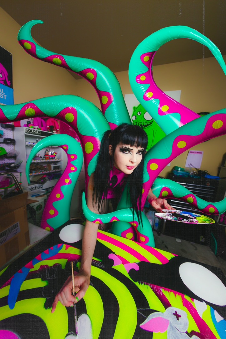 Person with black hair and makeup surrounded by colorful, tentacle-like sculptures in a room with shelves and boxes.