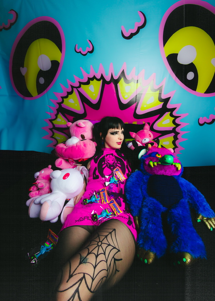 Person with black hair and pink dress surrounded by plush toys, sitting in front of a colorful cartoon face backdrop.
