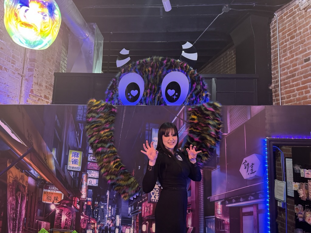 Person standing in front of colorful, large owl decoration with wide eyes, waving hands, in an indoor setting.