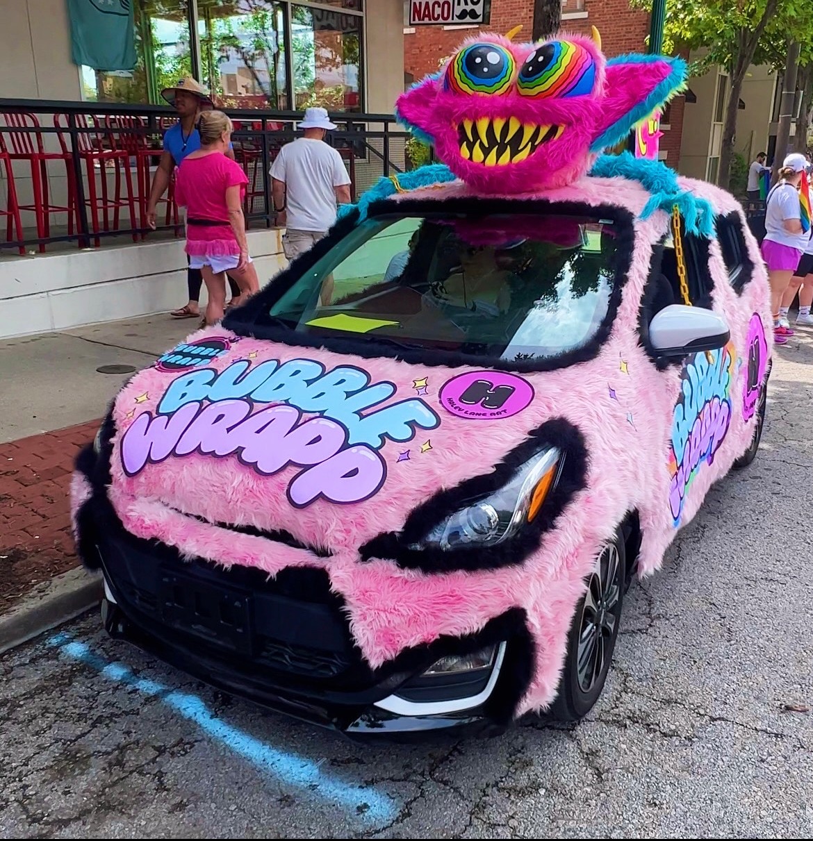 Pink car decorated with fuzzy material and a colorful monster head on top, parked on street with people nearby.