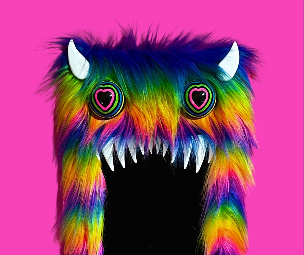 Colorful furry monster with heart-shaped eyes, white horns, and sharp teeth on a pink background.