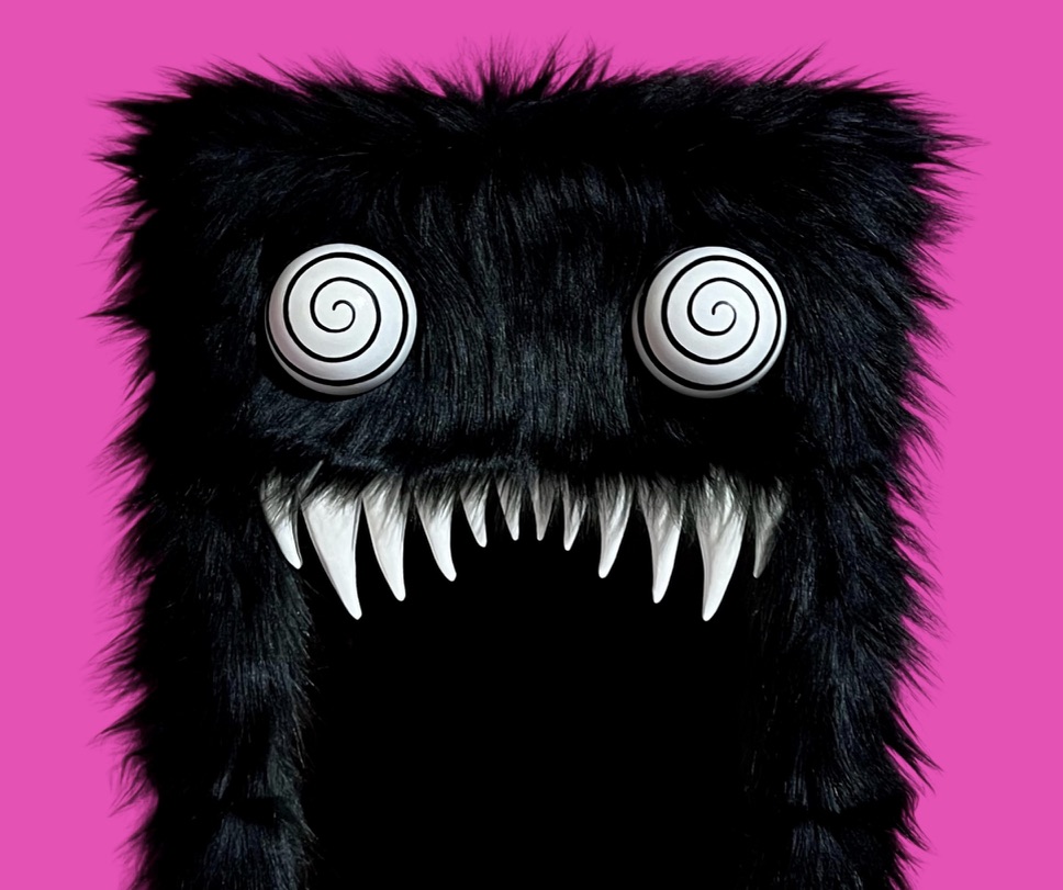 Black furry creature with spiral eyes and sharp teeth against a pink background.