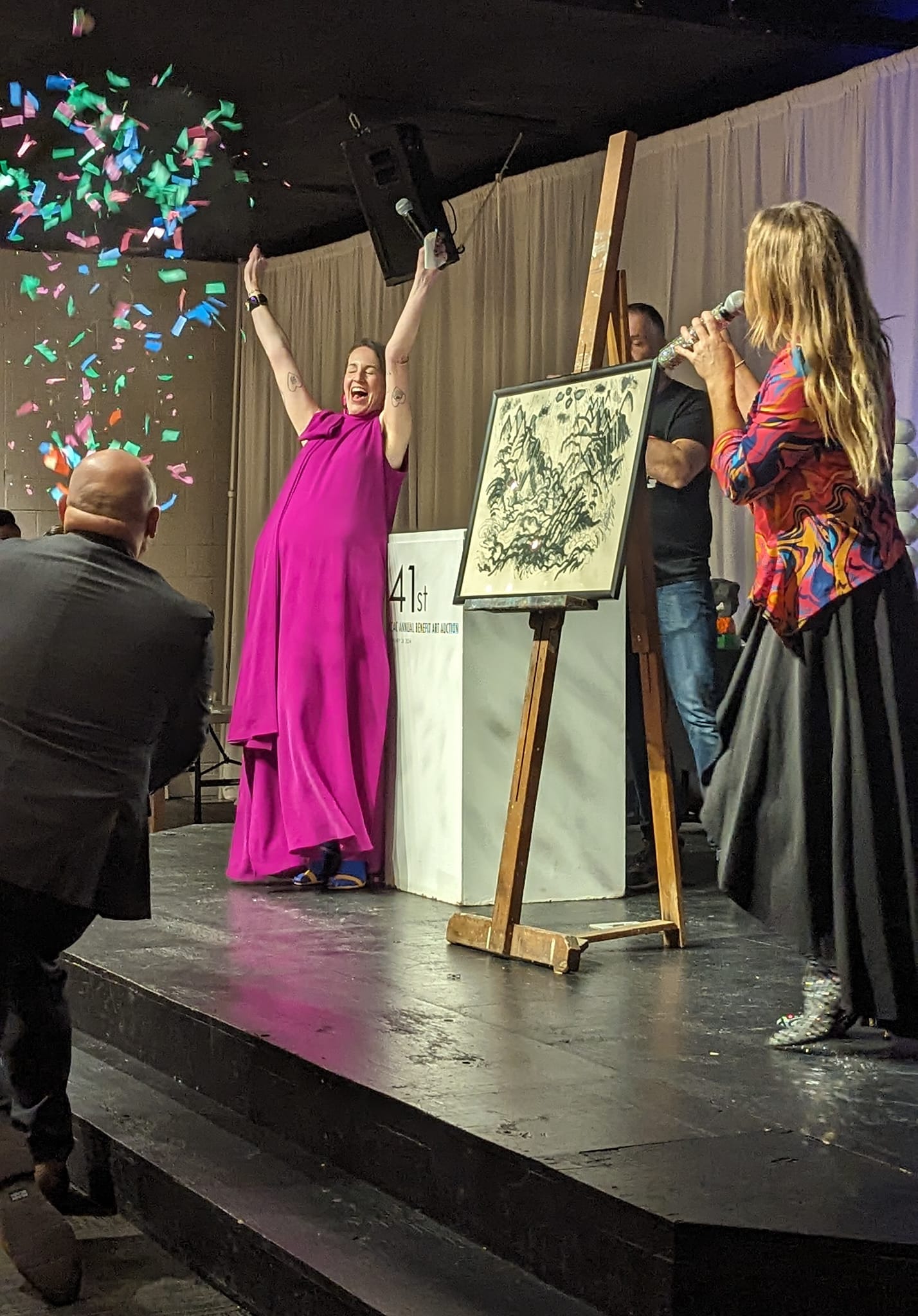 Woman in pink dress celebrates on stage with confetti, while another woman speaks into a microphone, and a man takes photos.