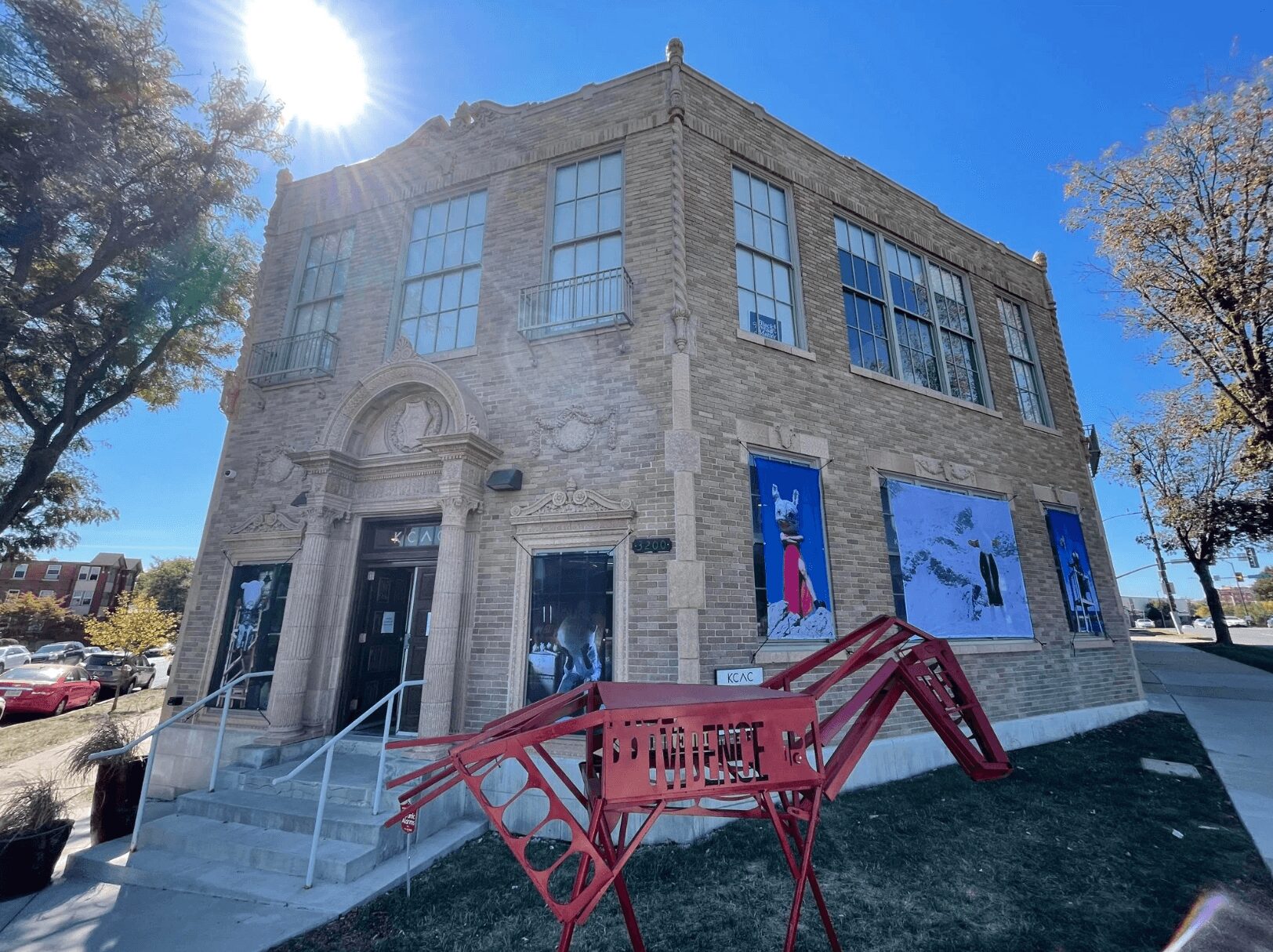 Corner of a historic brick building with large windows and banners, red sculpture in front, trees and blue sky in background.
