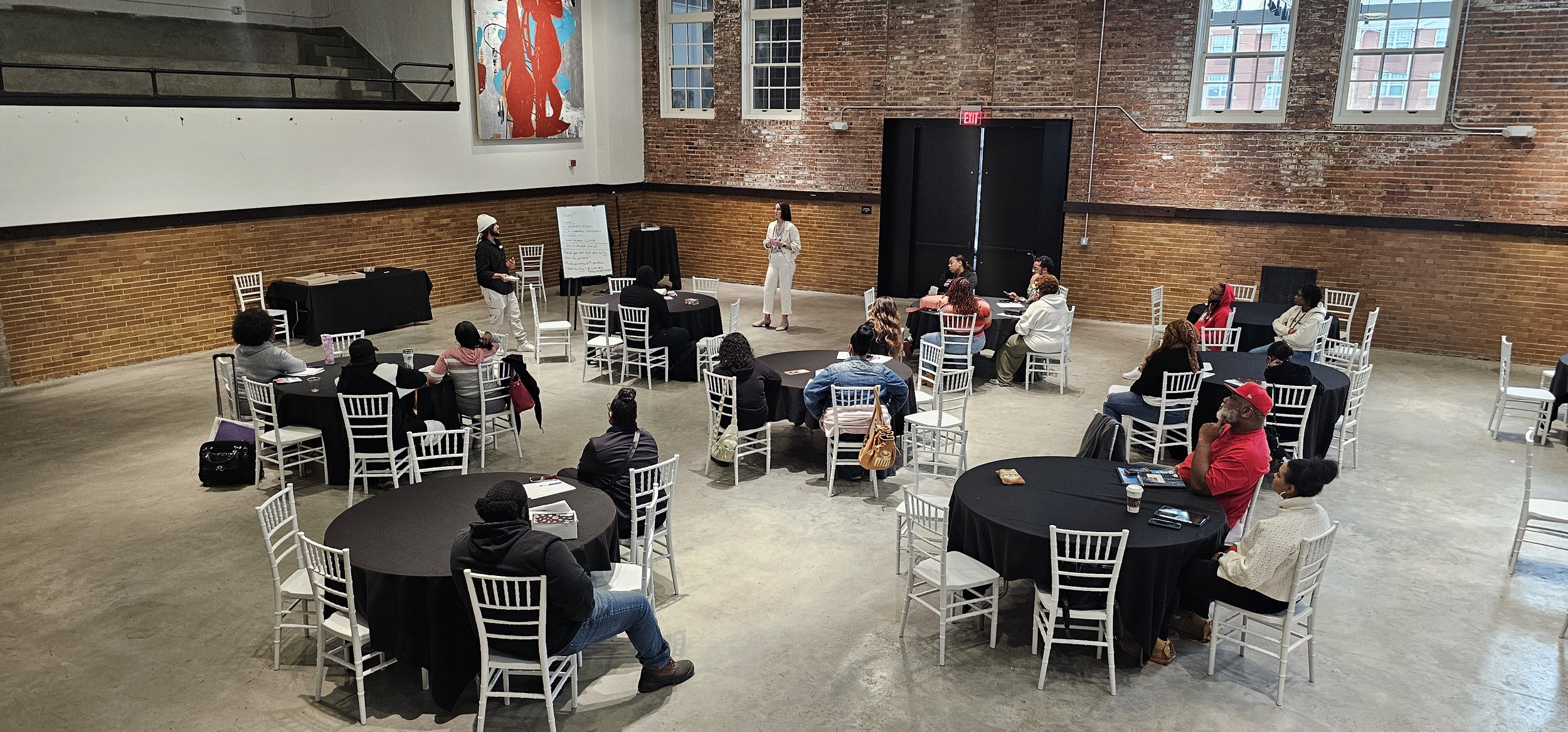 People seated at round tables in a spacious room with brick walls and large windows.