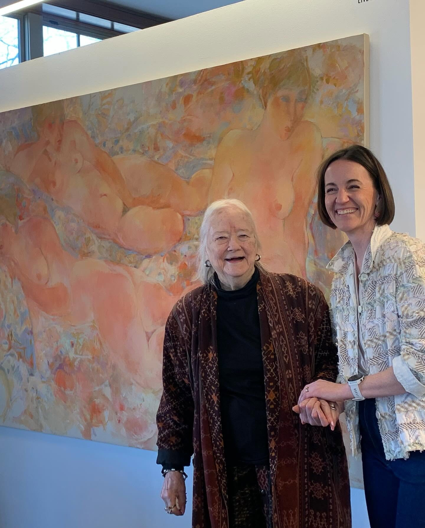 Two women smiling and holding hands in front of an abstract painting with warm colors.