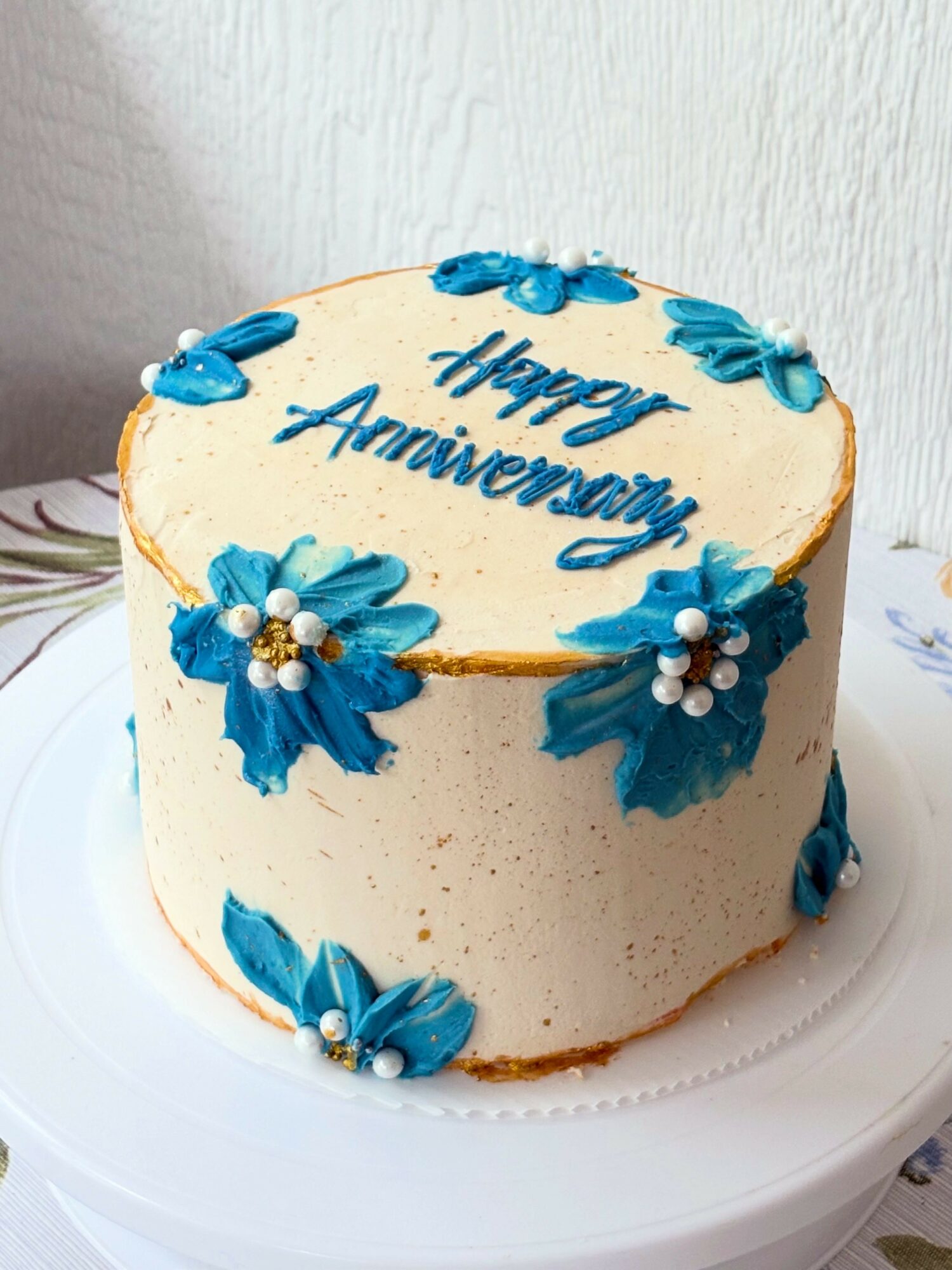 Round cake with white frosting, blue flowers, and a message for an anniversary, on a white cake stand.