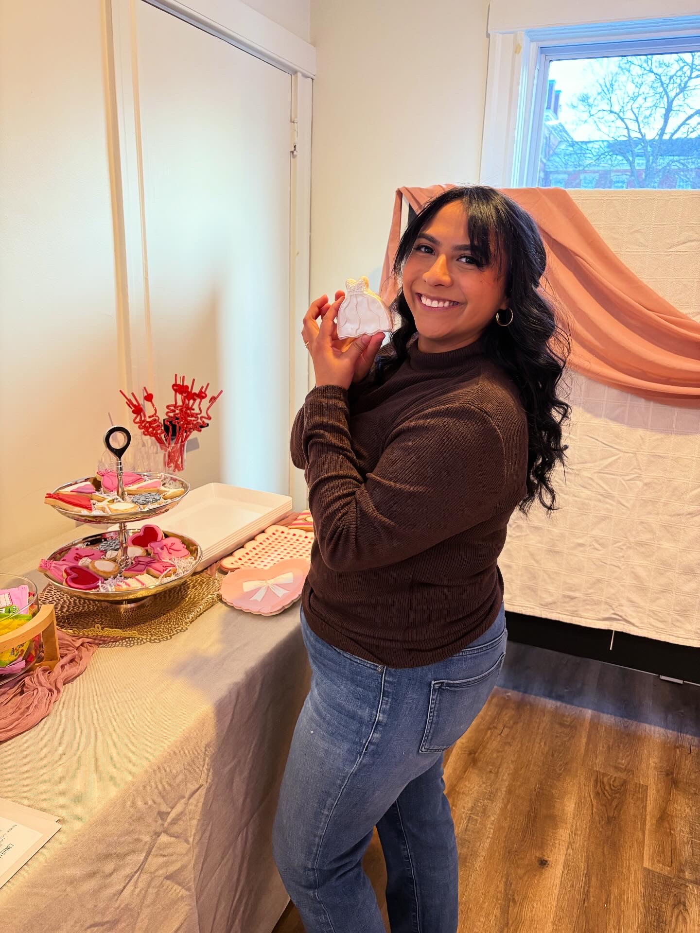Smiling woman with dark hair and bangs, wearing a brown top and jeans, standing indoors near a table with pink decorations.