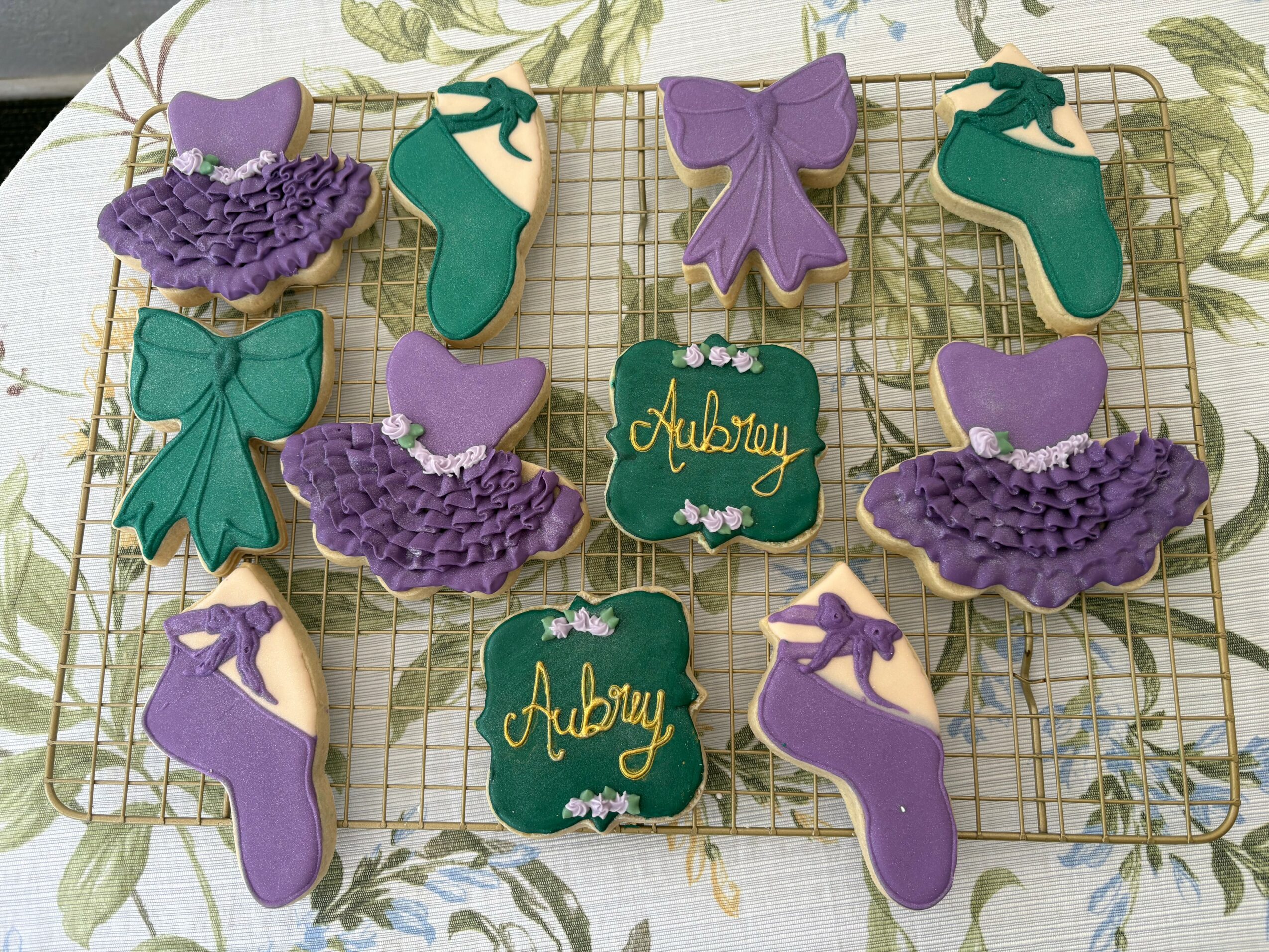 Decorative cookies shaped like ballet slippers, bows, and a sign reading 'Aubrey' in purple, green, and gold colors, arranged on a grid.