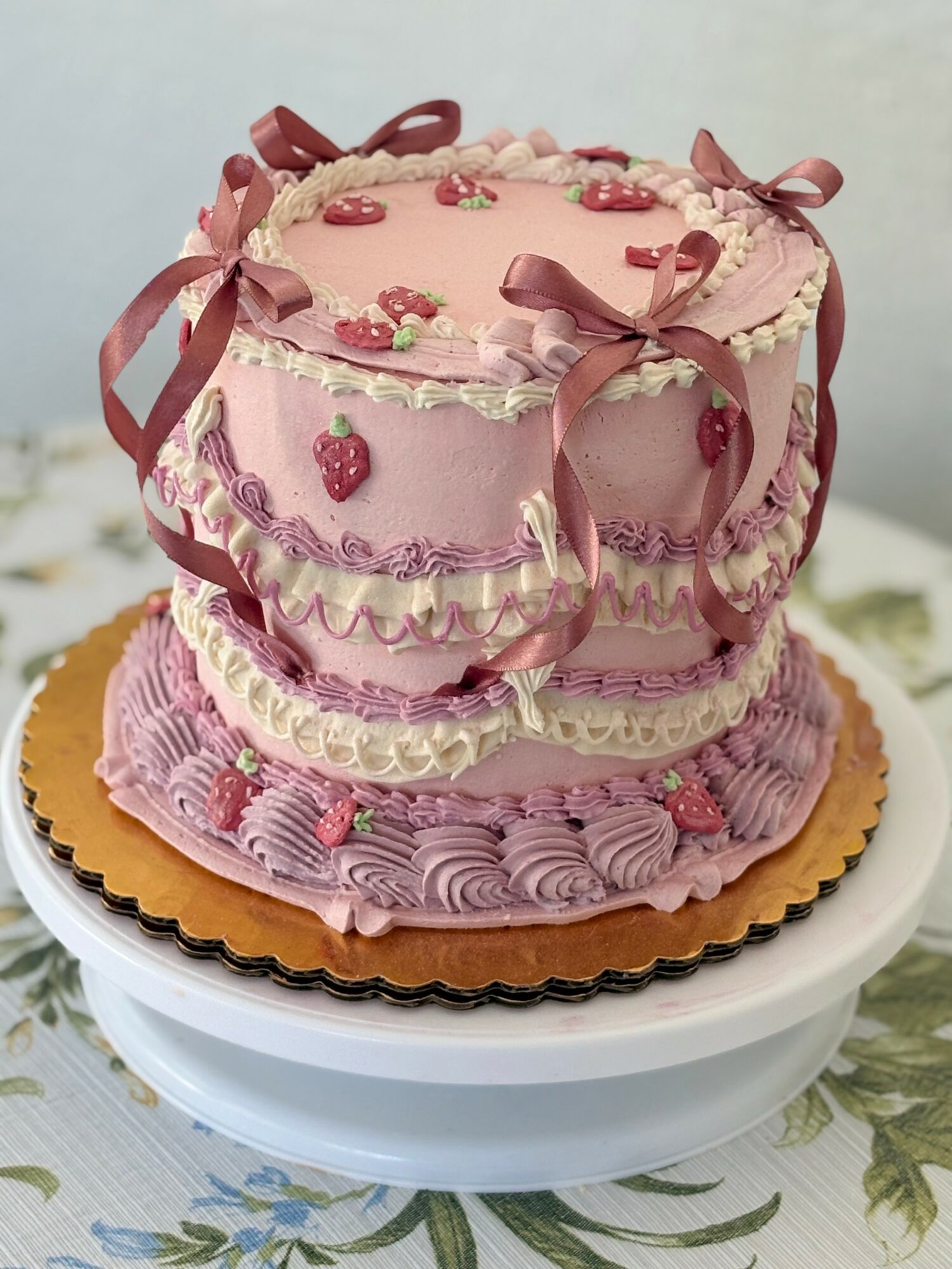Pink layered cake decorated with strawberries, ribbons, and ruffles, on a white cake stand with a floral tablecloth.