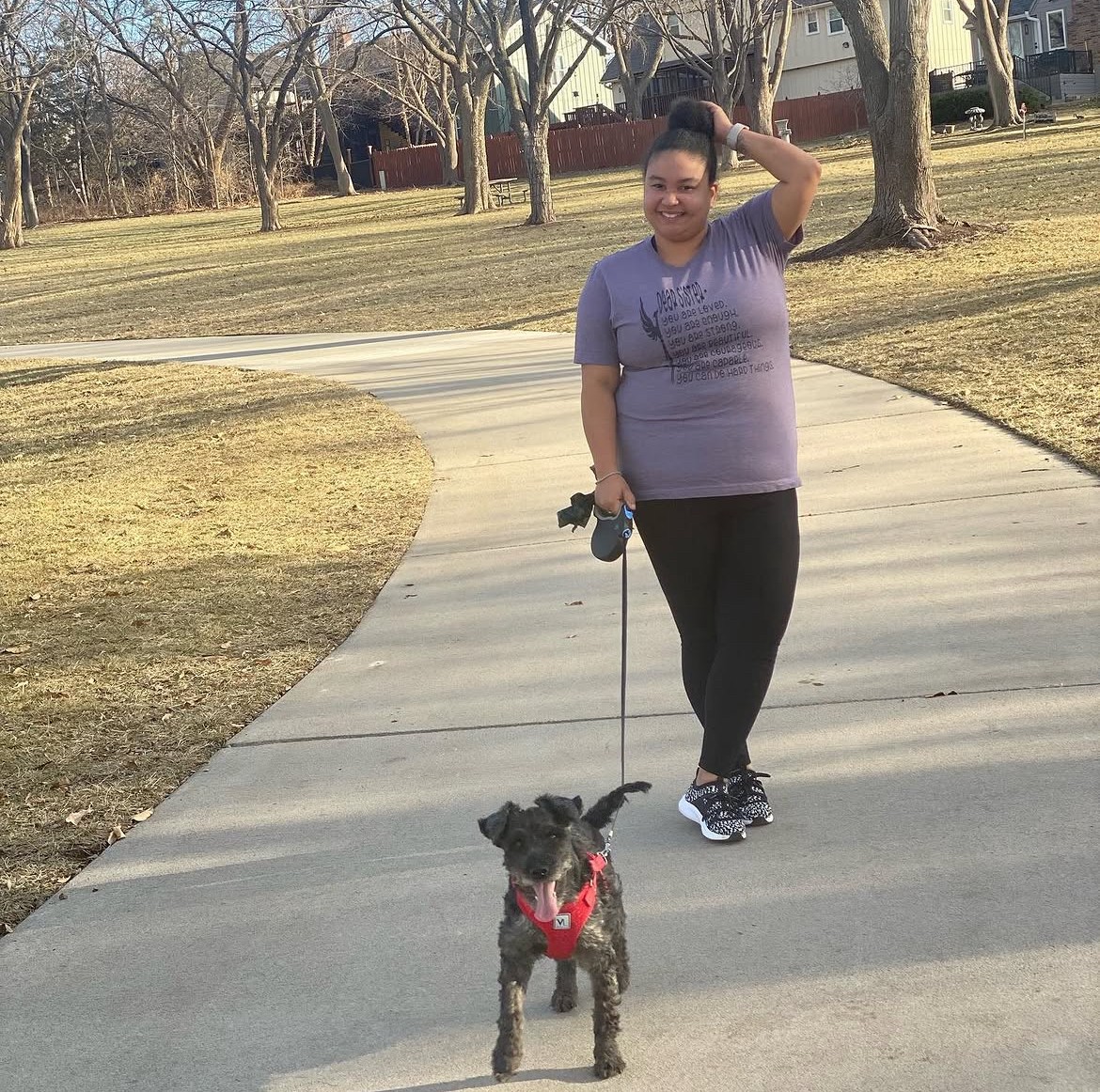 Person walking a dog on a curved sidewalk in a park with trees and grass, person smiling and holding the leash.