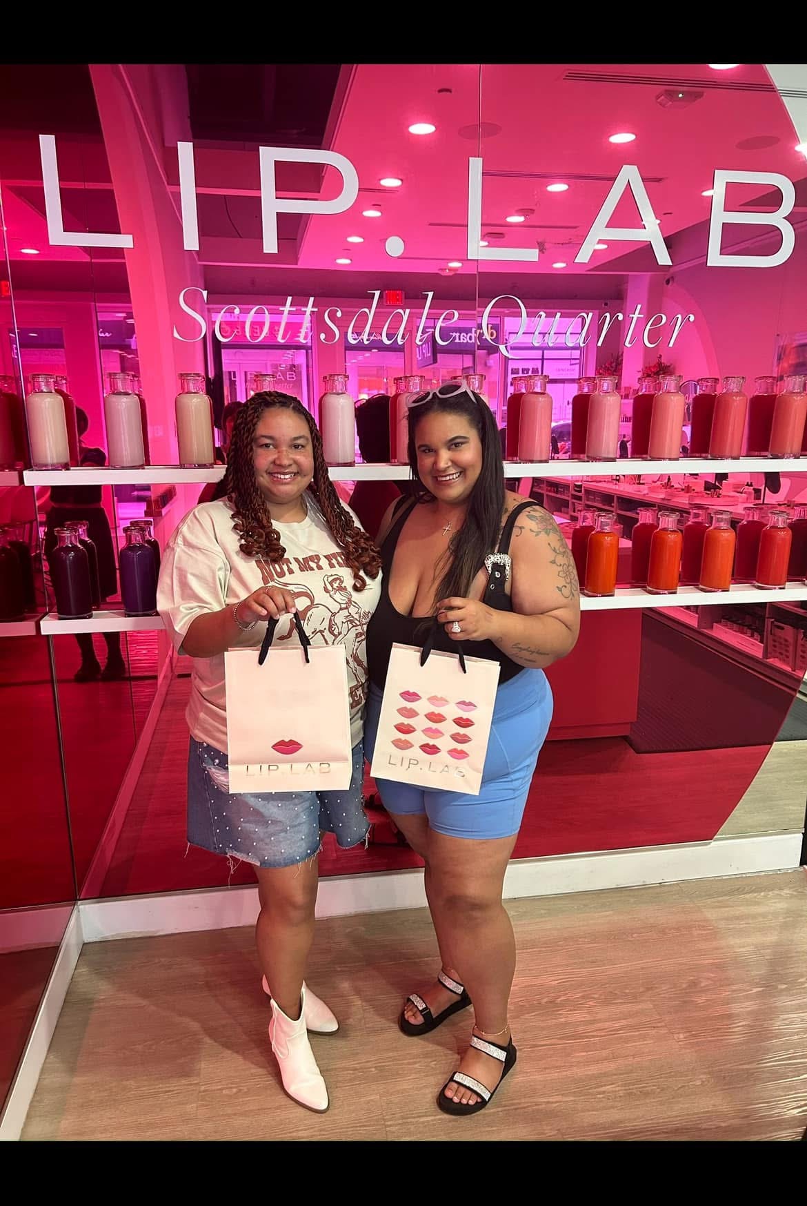 Two women stand in front of a pink Lip Lab store, holding shopping bags and smiling.