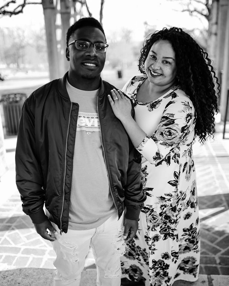 Two people standing outdoors, smiling, with trees and a walkway in the background.