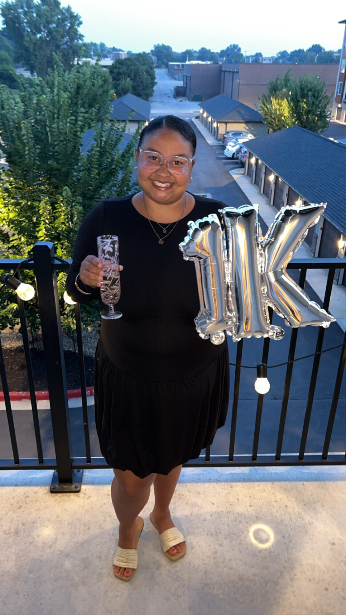 Woman smiling holding a drink and silver balloons spelling 'JIK' on balcony, evening background with trees and buildings.