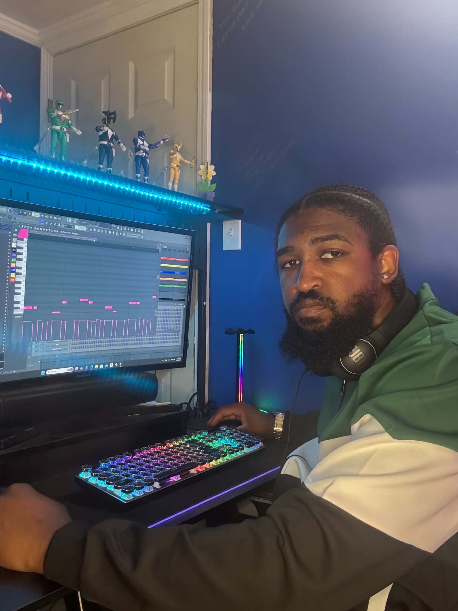 Person with headphones at a computer with a music production program, colorful keyboard, and blue ambient lighting.