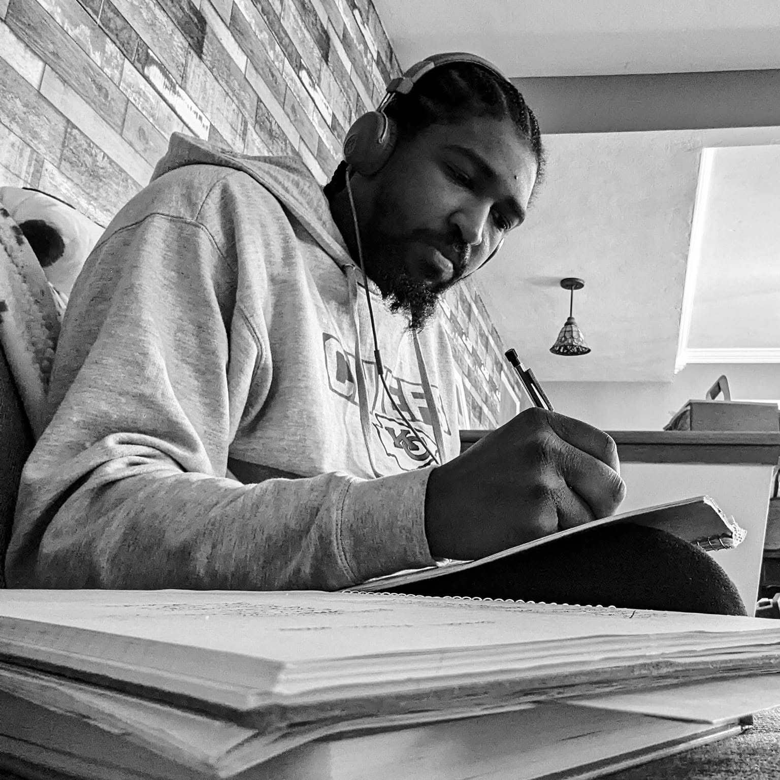 Man with headphones writing in a notebook, with a stack of papers in front, in a room with brick wall and ceiling light.
