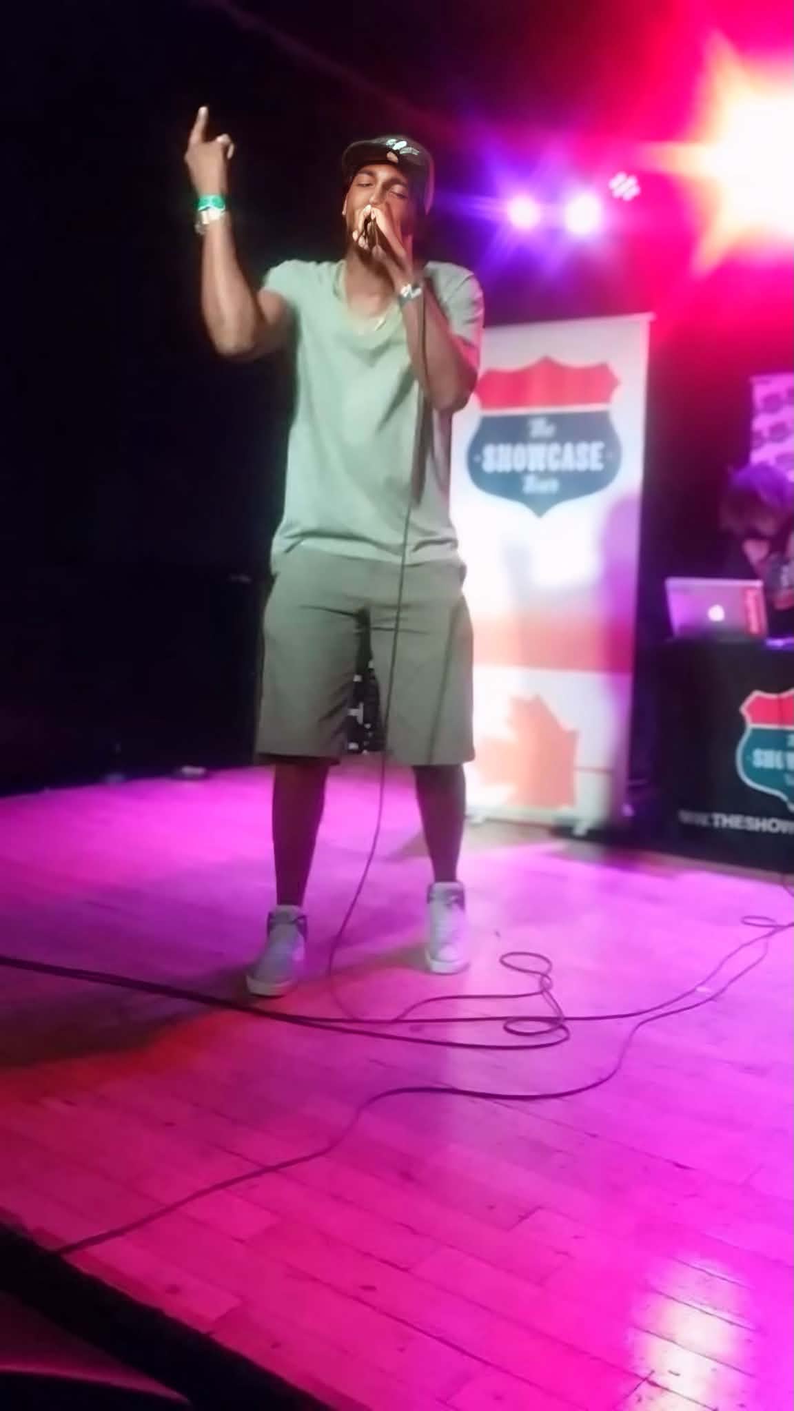 Person on stage holding microphone, wearing shorts and a t-shirt, with colorful stage lights in background.