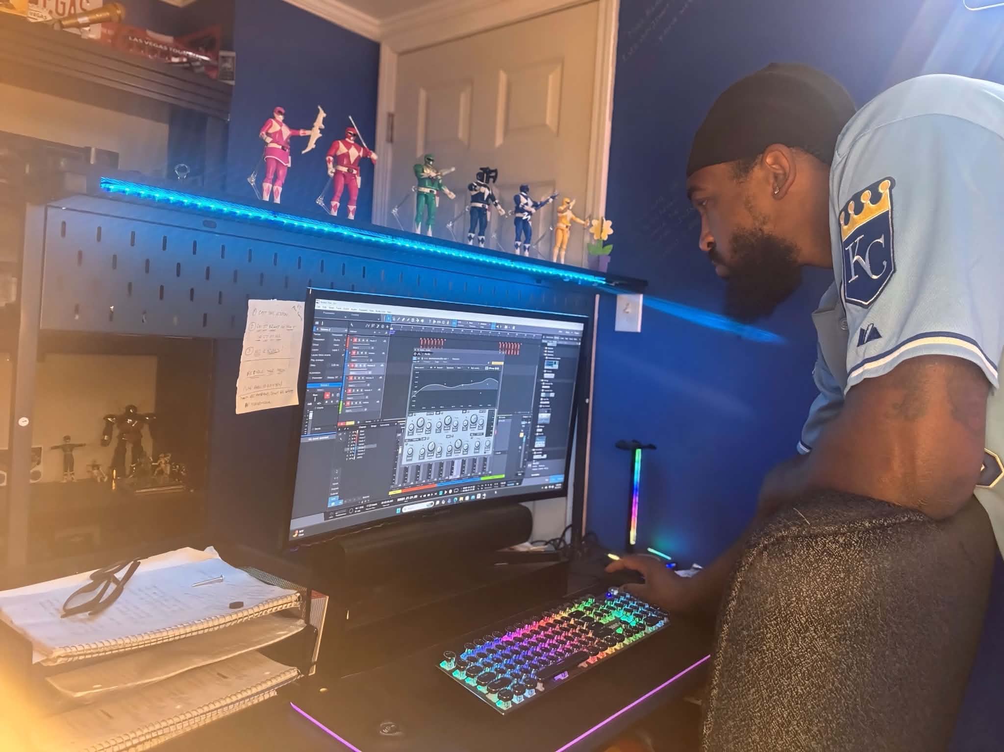 Person wearing a light blue shirt and black cap working on a computer with a colorful keyboard, shelves with toys and figures in background.