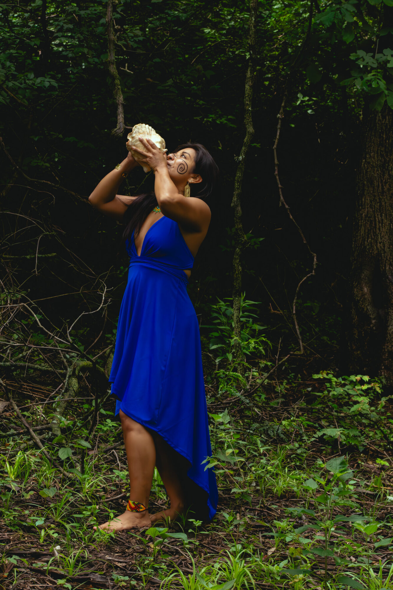 Woman in a blue dress holding a shell or object to her face in a forest setting with trees and greenery