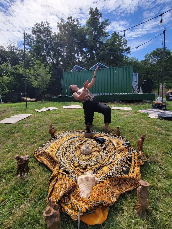 Person performing a dance move outdoors on grass, with a large patterned cloth on the ground, surrounded by small wooden stumps, trees, and a blue sky with clouds.