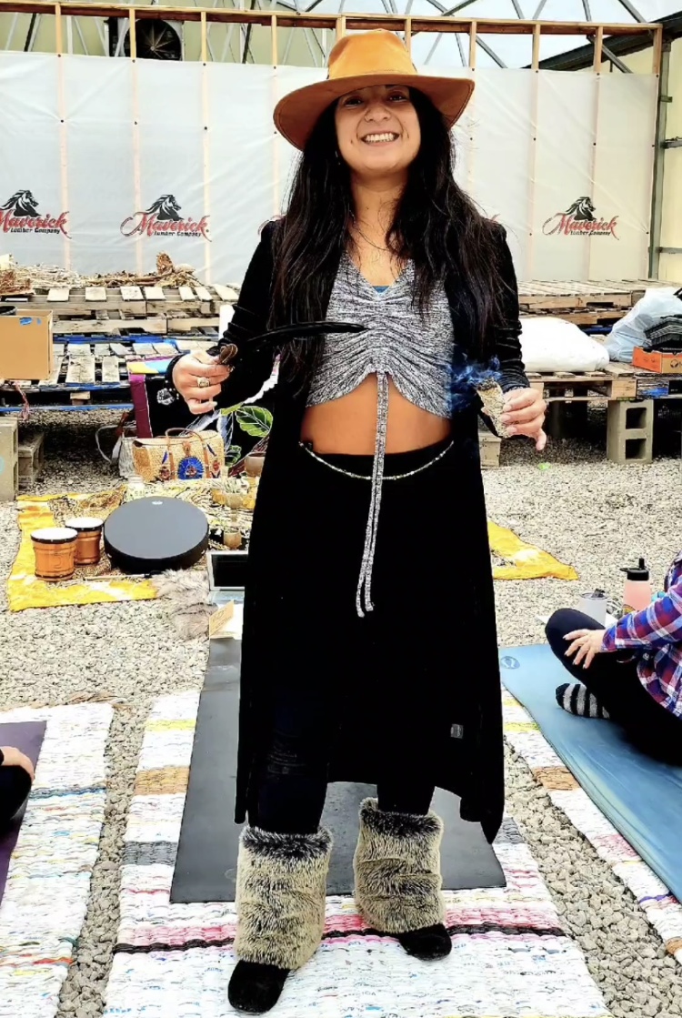 Woman with long dark hair wearing a wide-brimmed hat, smiling, standing outdoors with various items behind her.