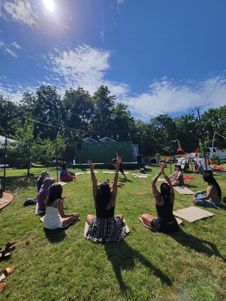 Group of people practicing yoga outdoors on a sunny day, sitting on mats with arms raised, surrounded by trees and blue sky.
