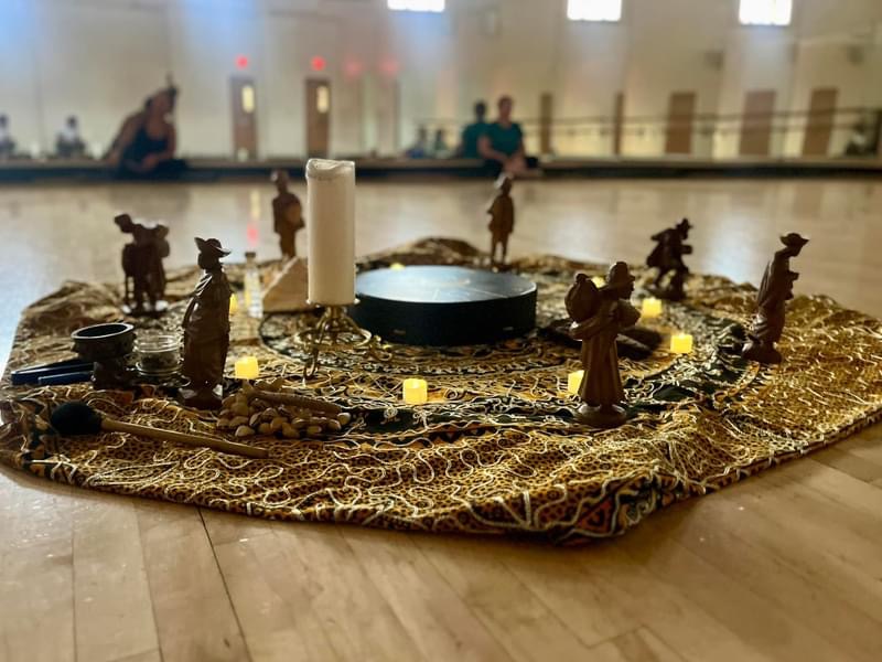 Decorative nativity scene with figurines, candles, and a central candle on a fabric cloth, set on a wooden floor indoors.