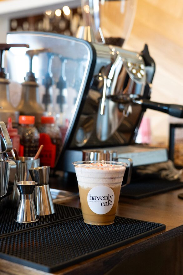 A plastic cup with a coffee drink and foam, labeled 'Havenly Cafe,' on a black mat at a coffee station.