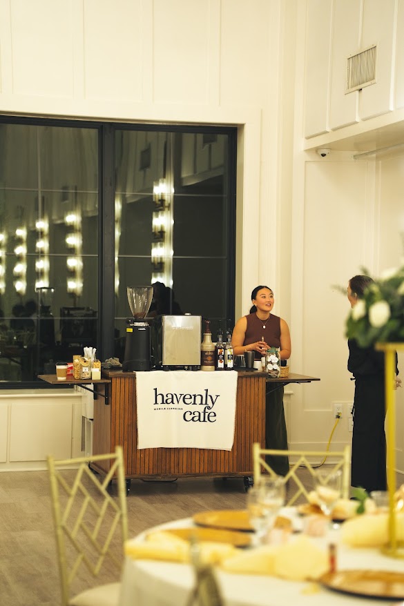 A woman stands behind a cafe counter with a sign reading 'heavenly cafe,' inside a well-lit room with large windows.