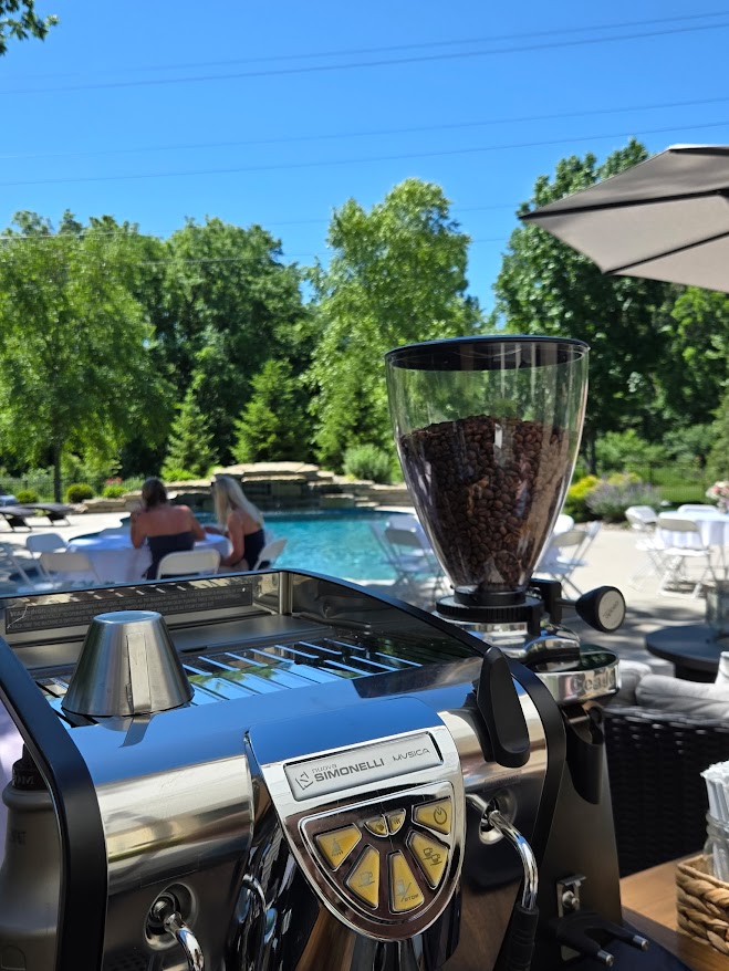 Coffee grinder and espresso machine outdoors near a pool with trees and people in the background.