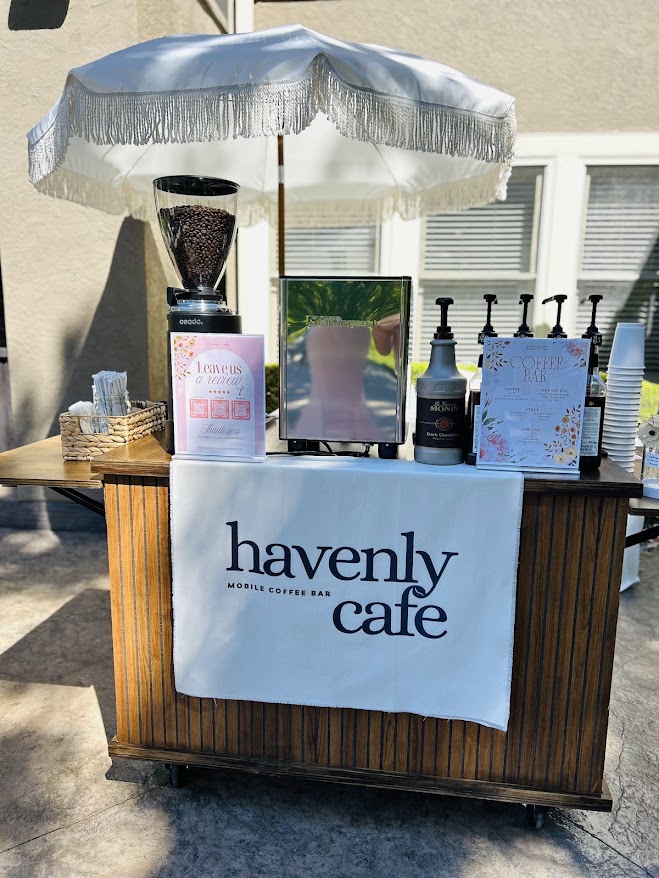 Coffee stand with a white umbrella, coffee beans, and drinks, featuring a 'heavenly cafe' banner, outdoors on a sunny day.