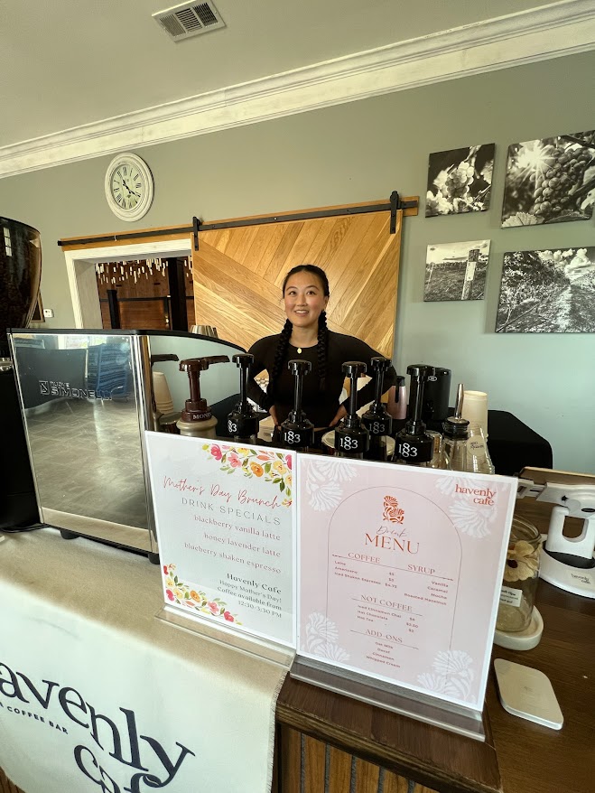 Woman standing behind a counter with menu signs, coffee equipment, and a computer in a cafe setting.