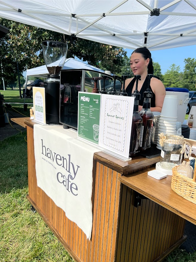 A woman behind a cafe counter under a white canopy, with menus and coffee equipment, outdoors on a sunny day.