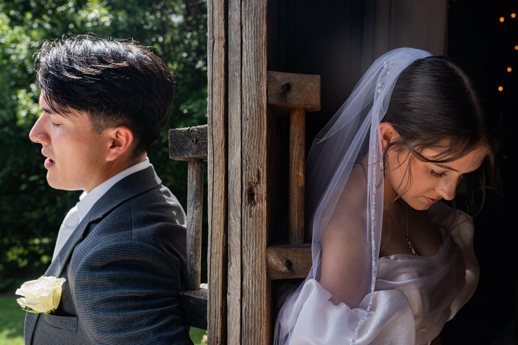 A bride and groom stand back to back, separated by a wooden structure, with the bride looking down and the groom outdoors.
