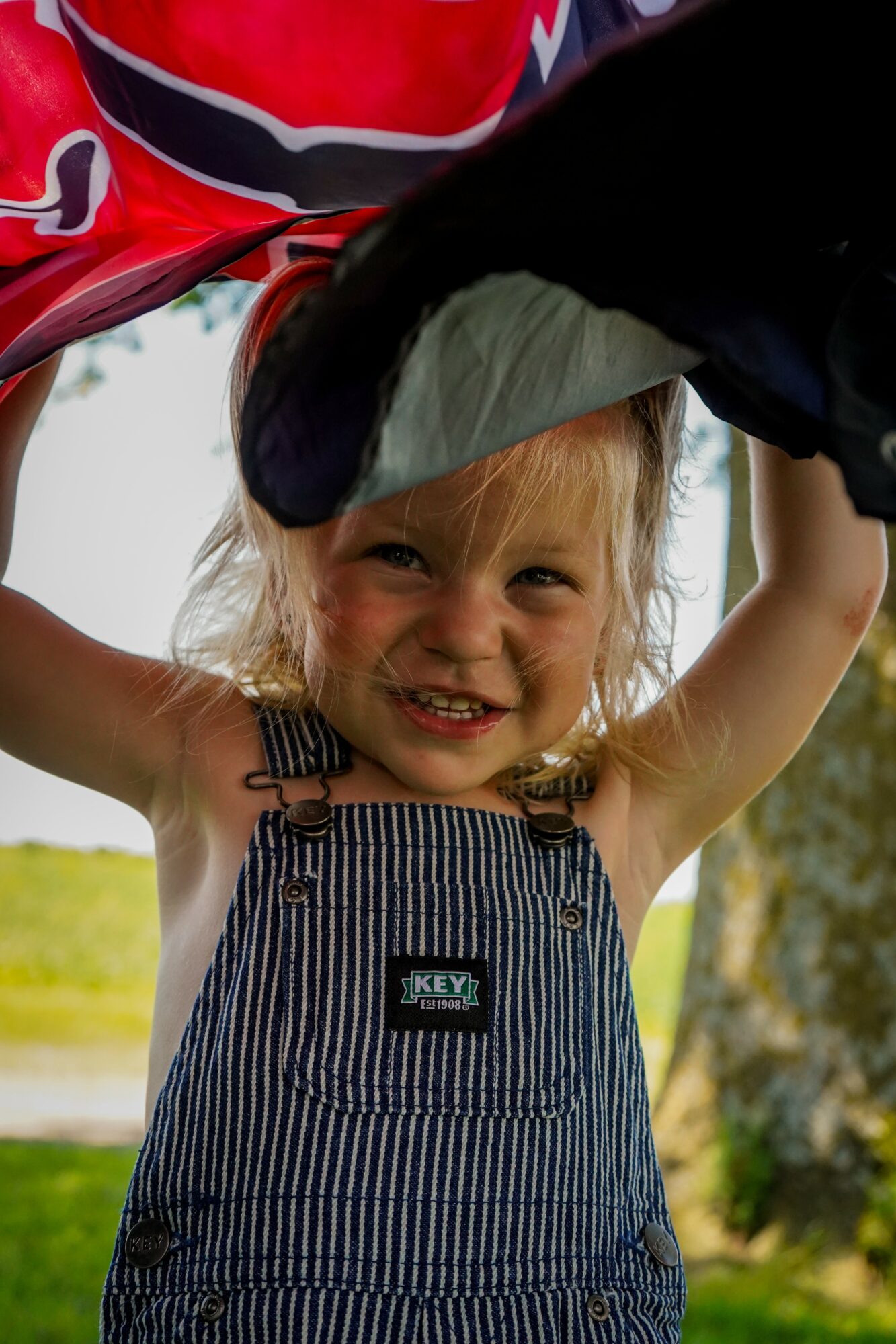 Young girl outdoors, smiling, holding a black object above her head, wearing striped overalls and a green hat, trees in background.