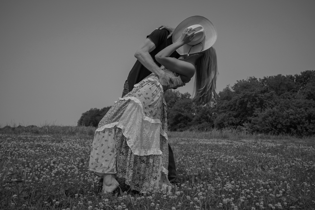 Two women in a field, one bending and the other standing, with one holding a hat over the other's head.