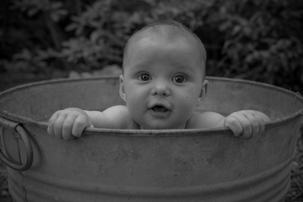 Baby with wide eyes and open mouth holding onto the edge of a round container outdoors, background of foliage