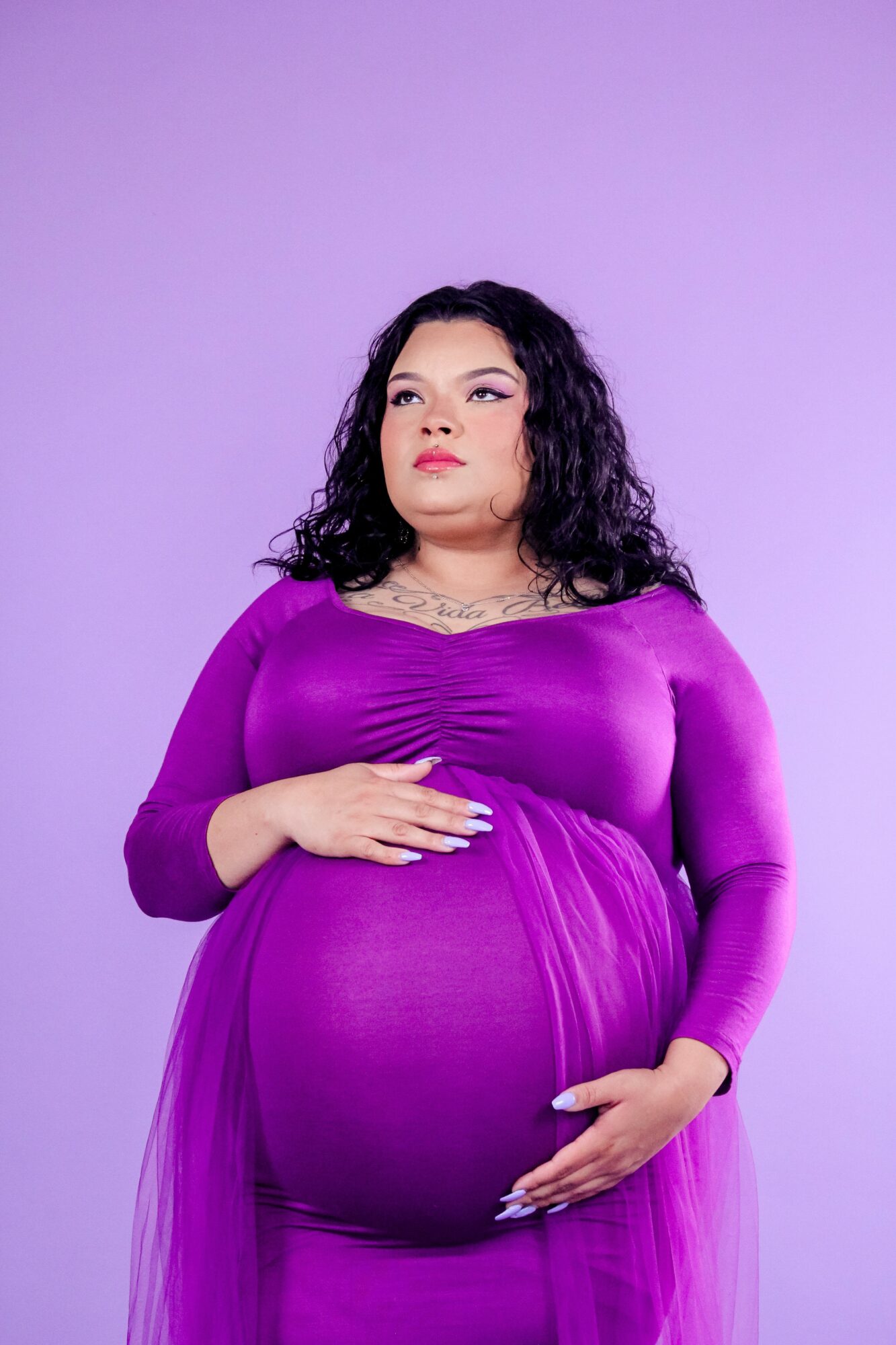 Pregnant woman with curly hair wearing a purple dress, standing against a purple background, holding her belly with both hands.