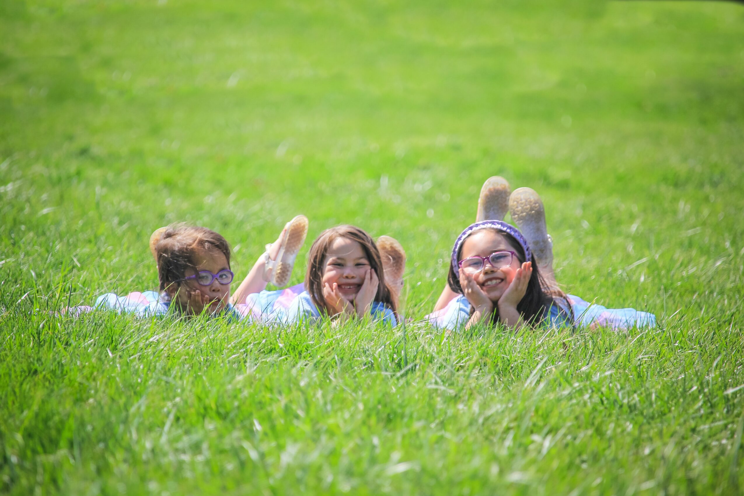 Three children lying on grass, smiling and looking at the camera, with a green field background.