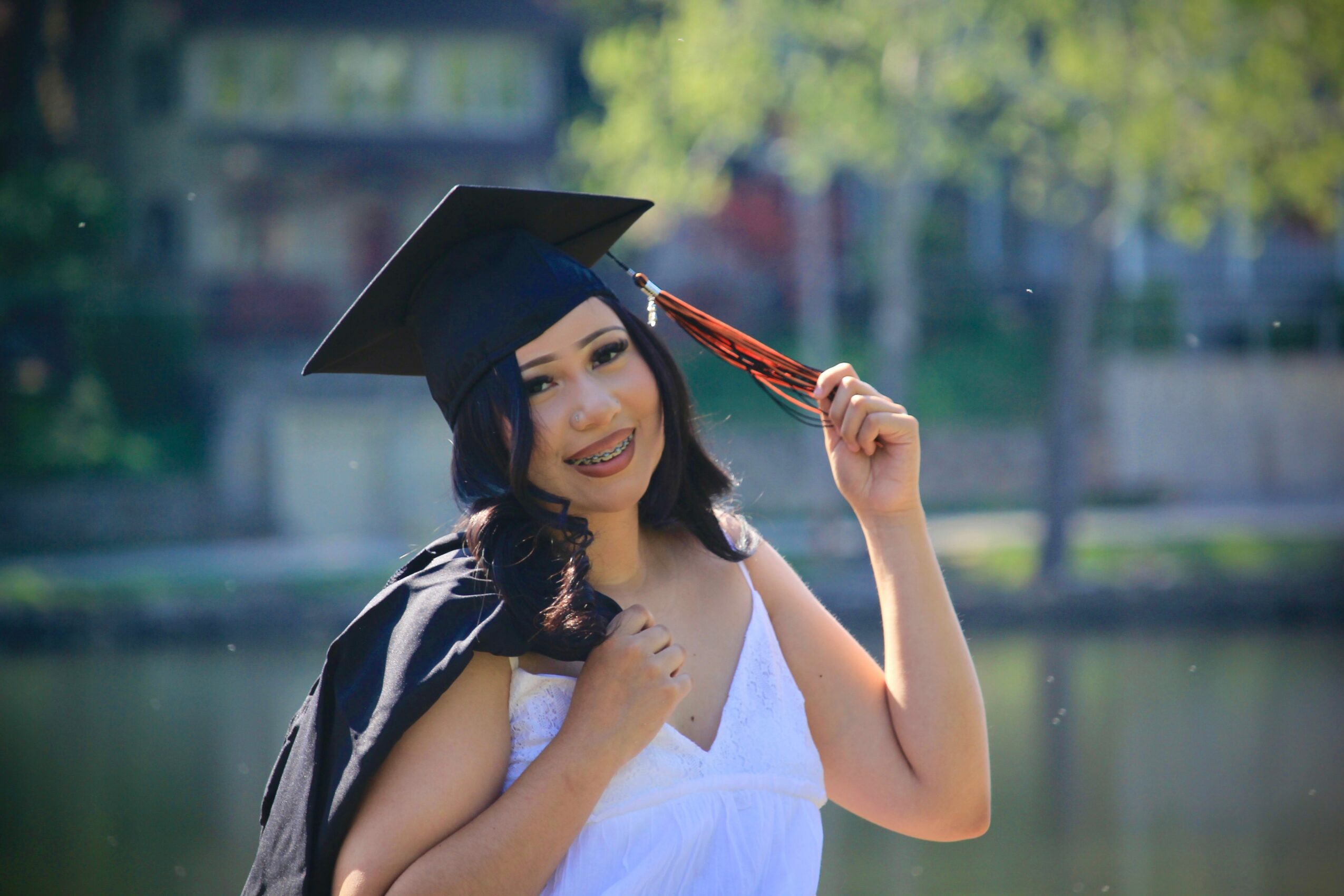 Young woman in graduation cap and gown smiling outdoors near water, holding her tassel.