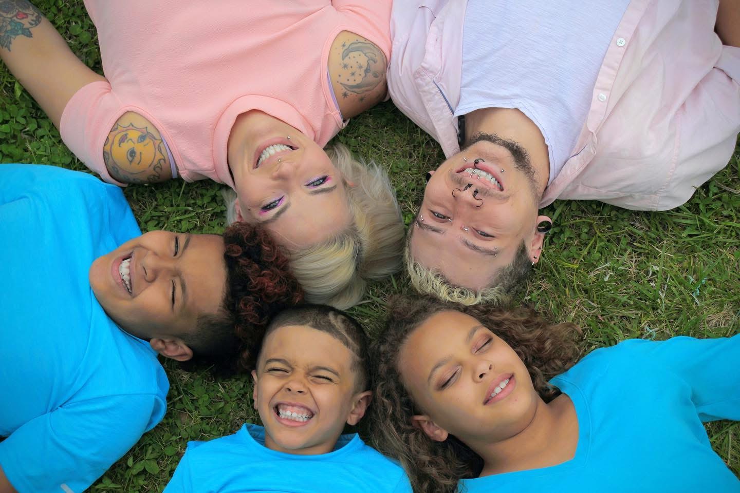 Five people lying on grass in a circle, smiling and looking up at the camera, heads close together.