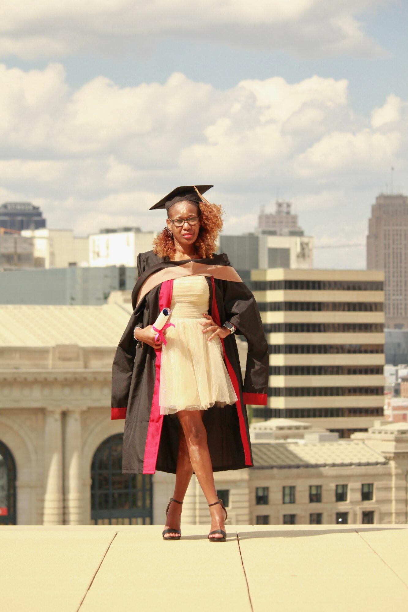 Person in graduation gown and cap standing outdoors with city buildings in background.