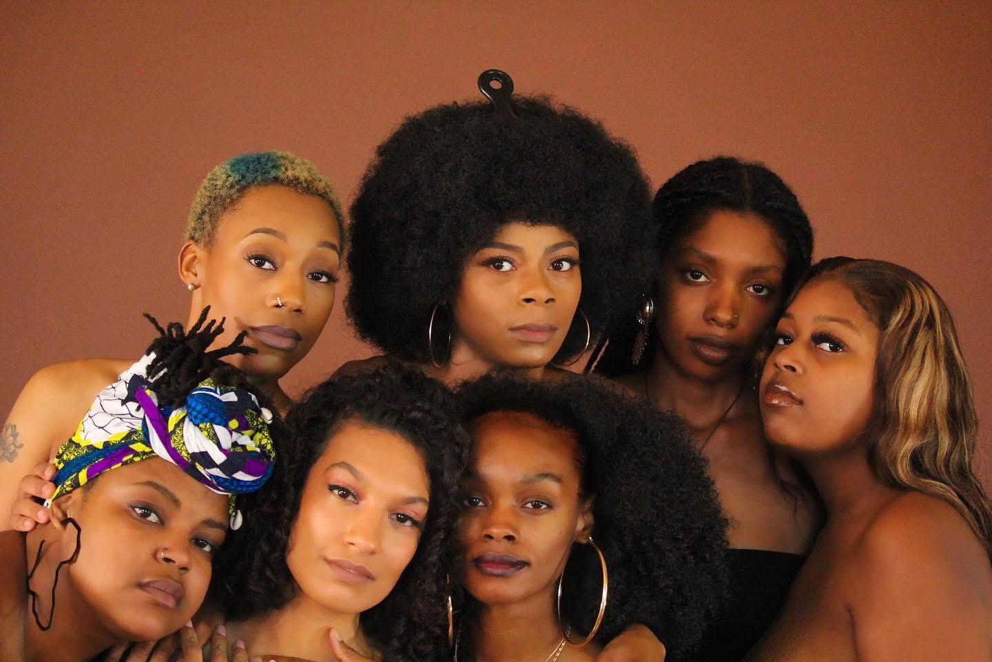 Group of seven women with diverse hairstyles and expressions posing together against a plain background.