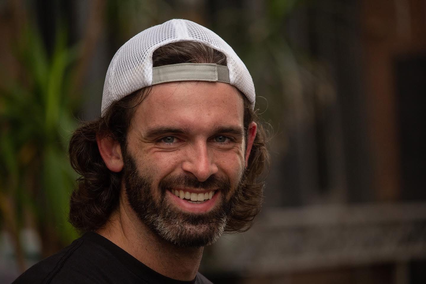 Man with long hair, beard, wearing a white cap backwards, smiling, in an outdoor setting with greenery.