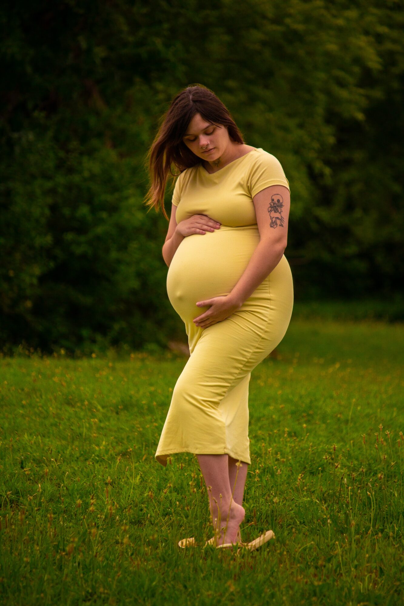 Pregnant woman in yellow dress standing outdoors on grass, holding her belly, with trees in background.