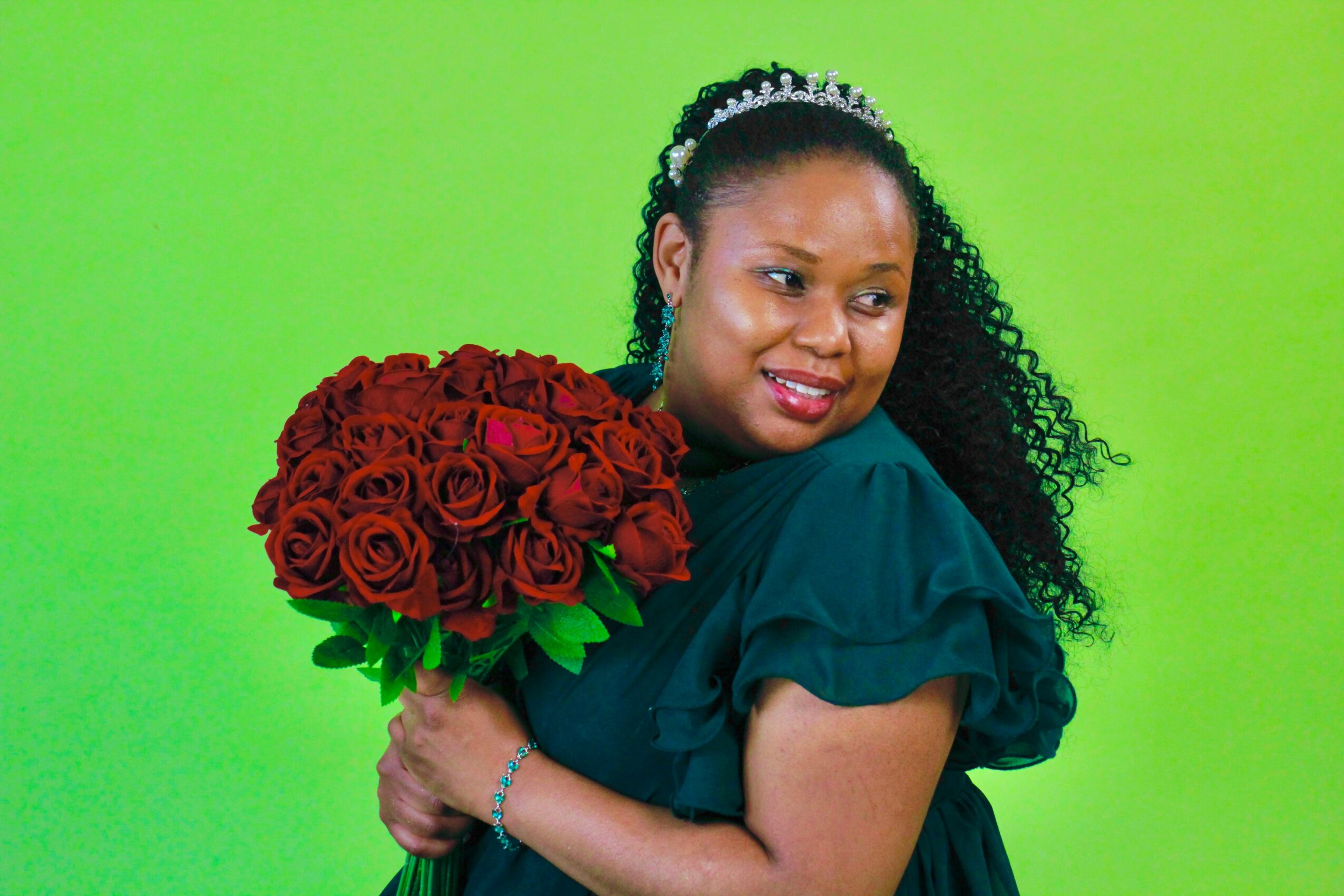 Woman with curly hair wearing a tiara and dark dress holding a large bouquet of red roses against a green background.