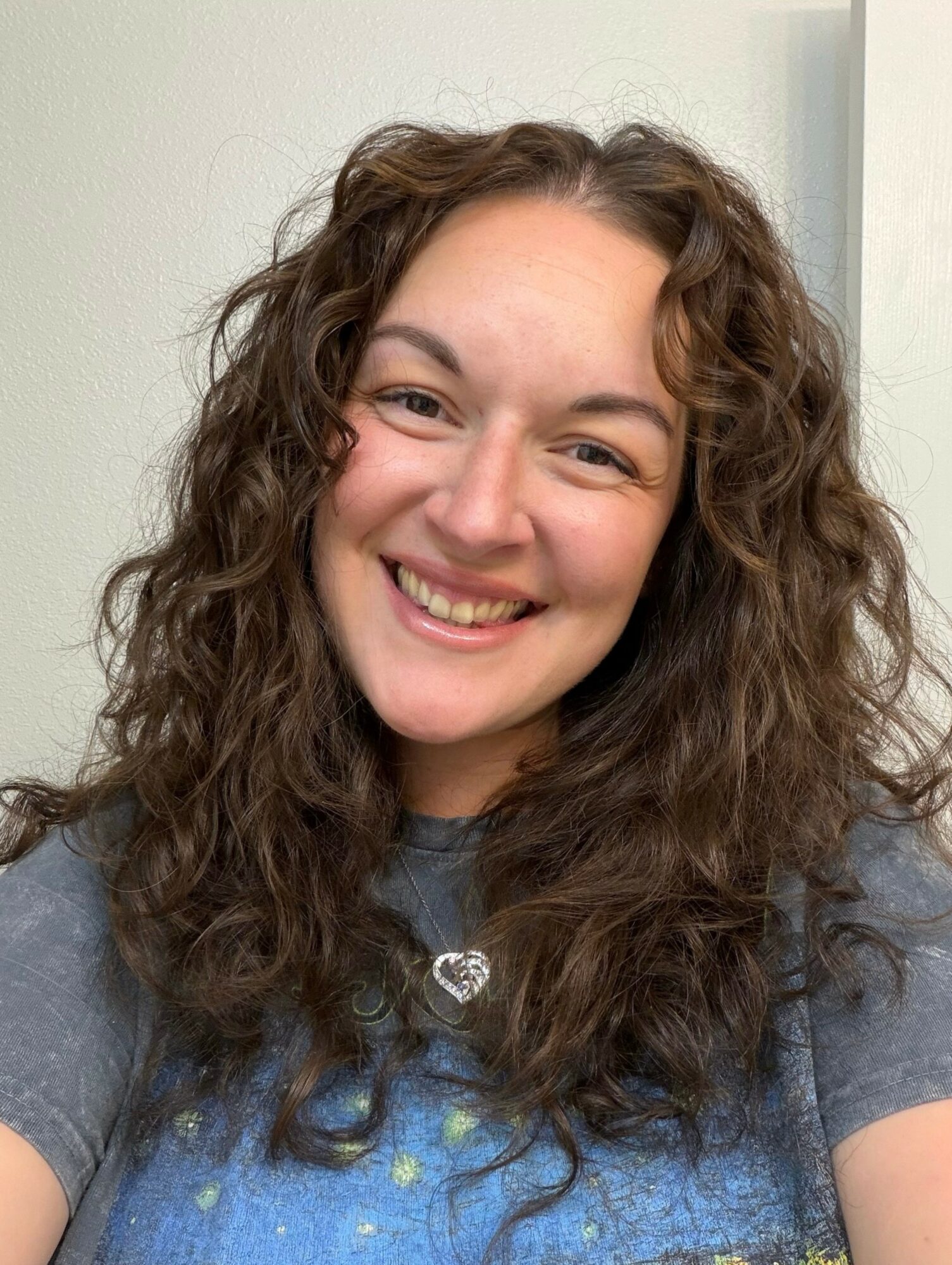 Young woman with curly hair smiling, wearing a gray shirt, standing indoors against a light background.